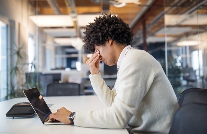 Een persoon achter een laptop, die zijn gezicht vasthoudt, oogt gestrest in een modern kantoor.