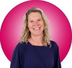 Vrouw met blond haar lacht in een donkerblauwe blouse, tegen een roze cirkelvormige achtergrond.