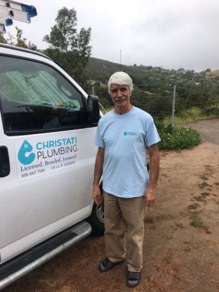 Man standing beside a white plumbing van on a dirt roadside, wearing a light blue shirt and tan pants.