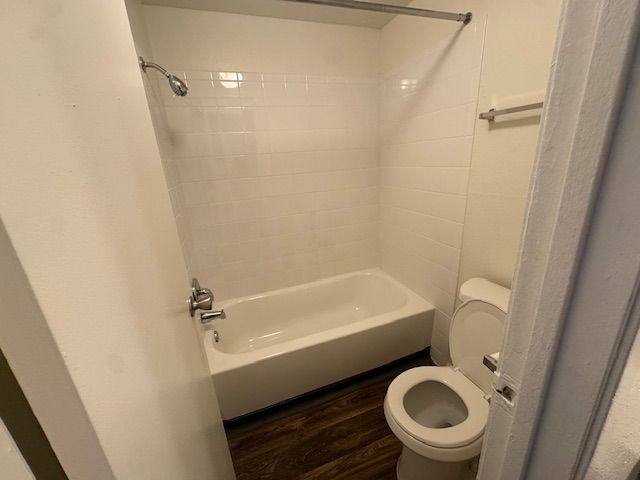 Small bathroom with a white tub, toilet, and shower curtain rod on dark wood flooring.