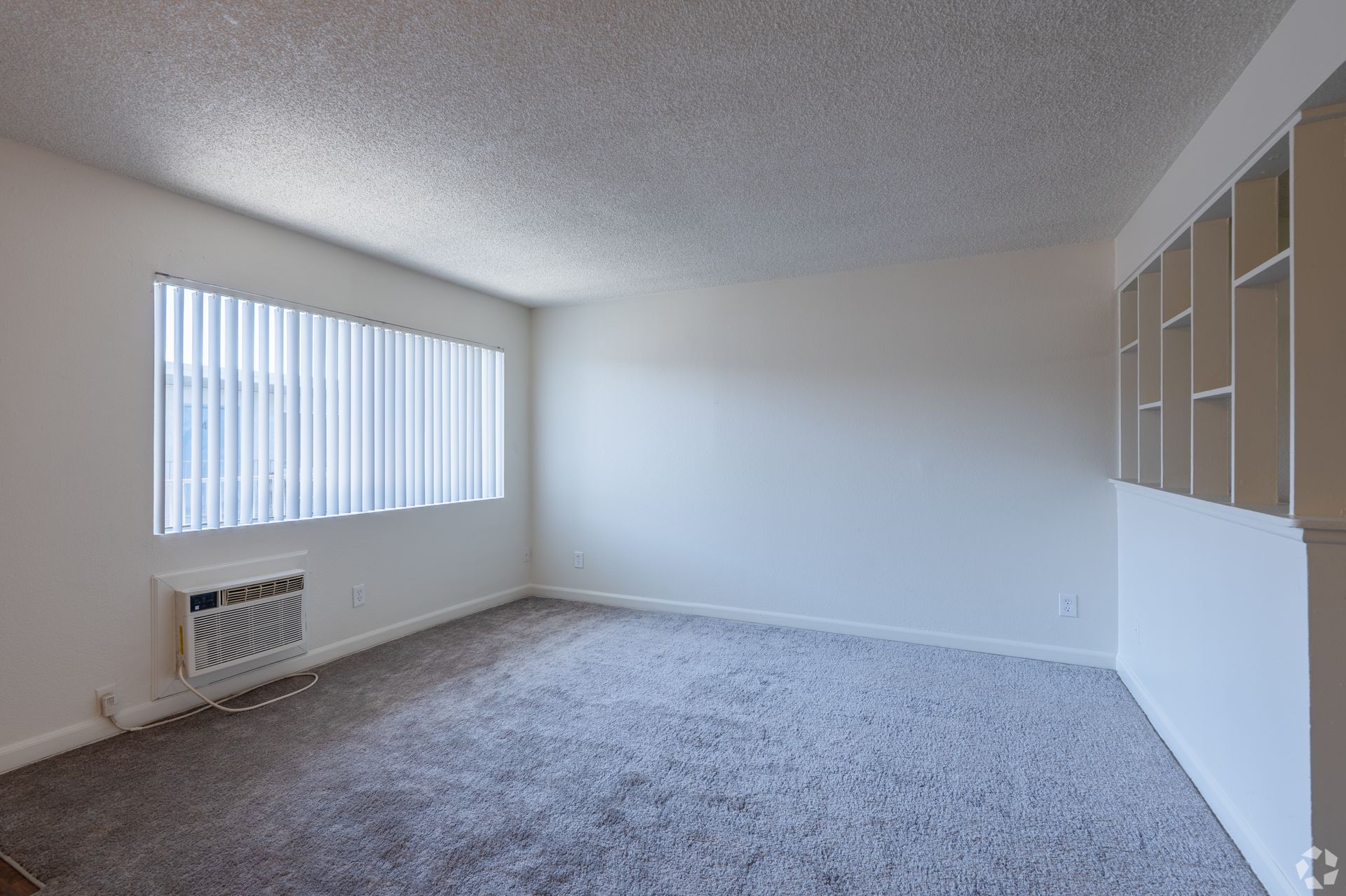 Empty carpeted room with white walls, a large window with blinds, and built-in shelving.