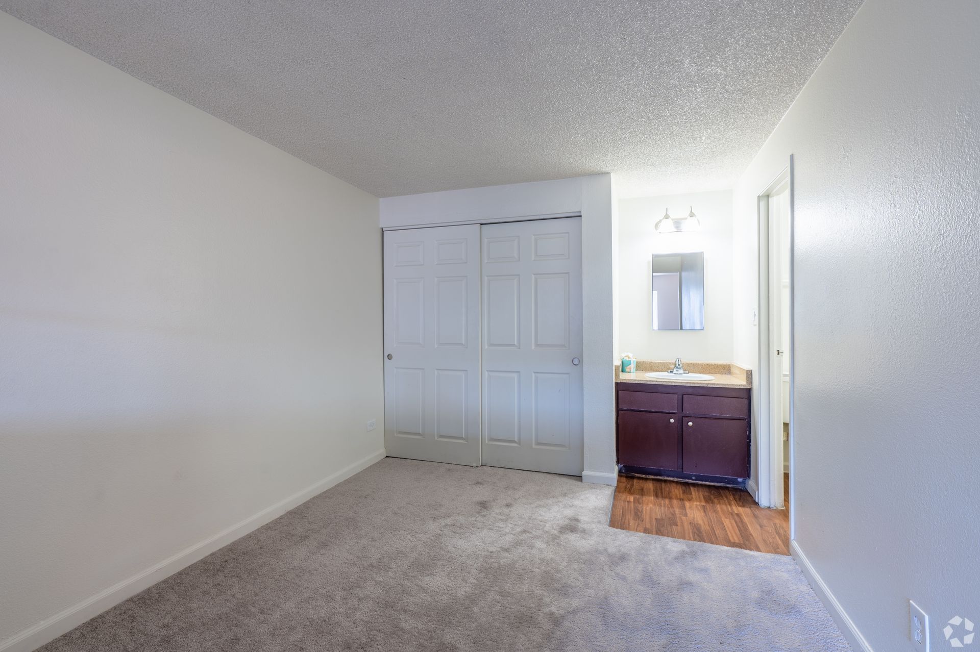 Empty bedroom with beige carpet, white walls, closet doors, and a small vanity near the door