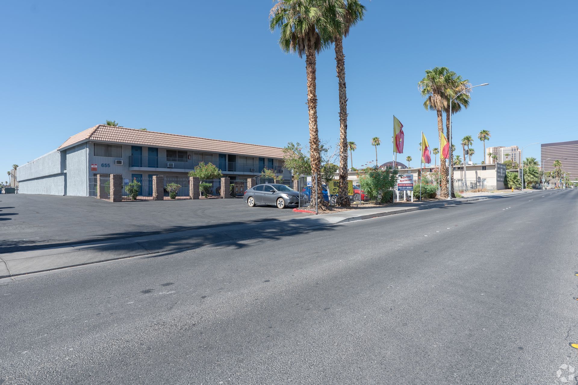 Wide street beside a low beige building with palm trees and parked cars under a clear blue sky
