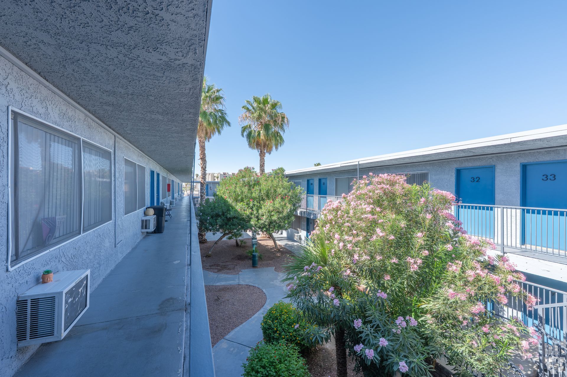 Apartment courtyard with palm trees, flowering shrubs, and blue-painted buildings under a clear sky