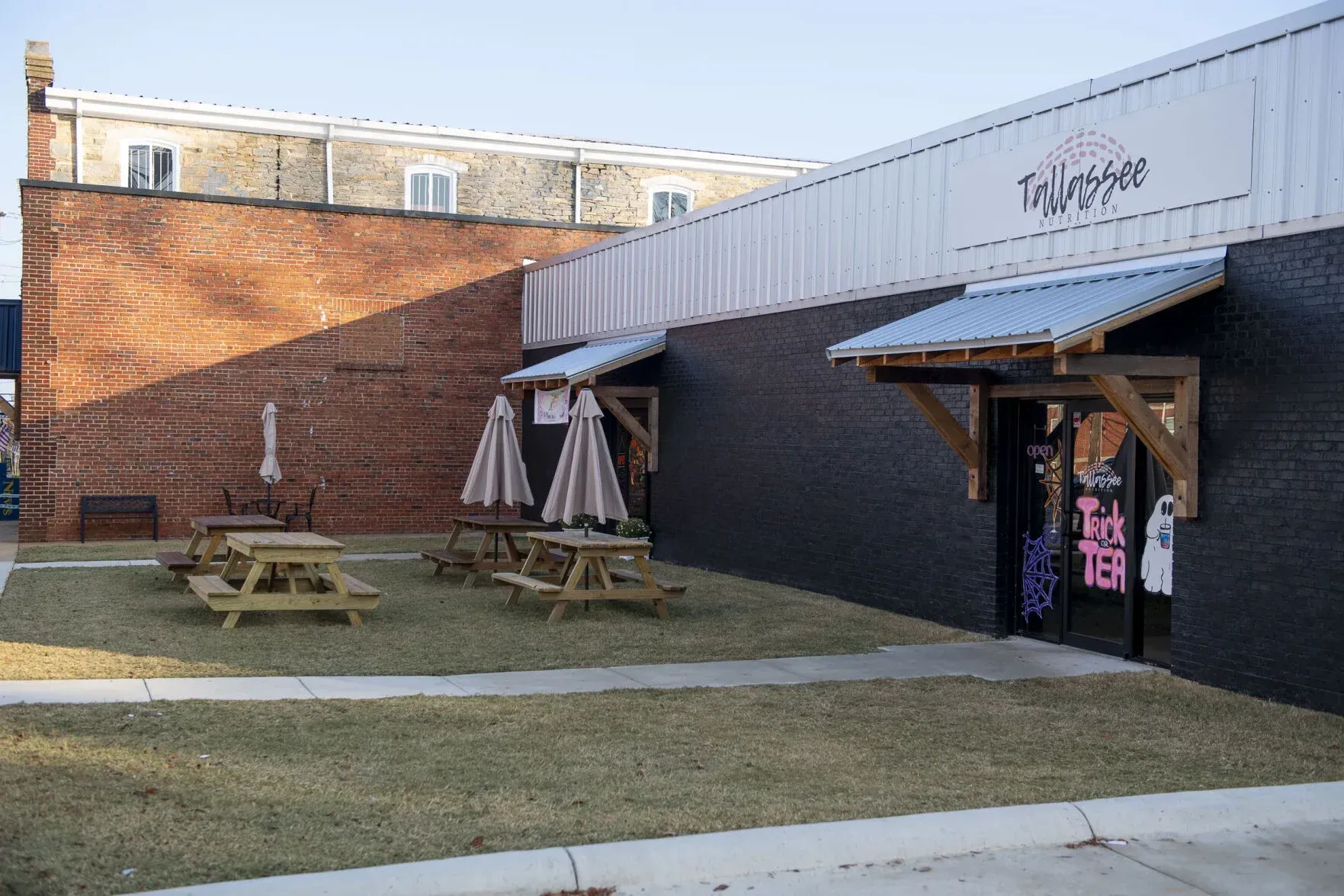 Exterior view of a business with a brick wall, black painted building, picnic tables, and awnings.