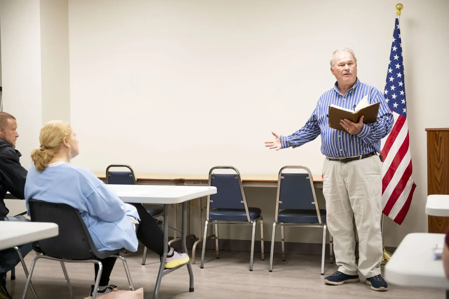 Man reading from a book, gesturing to a seated audience in a room with an American flag.