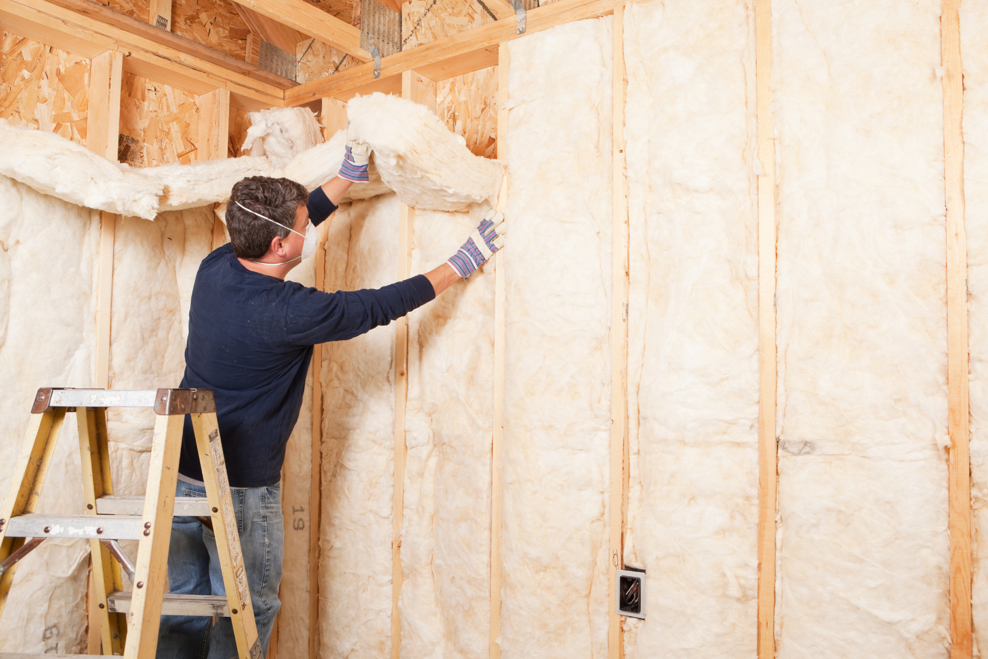 A man is standing on a ladder insulating a wall.