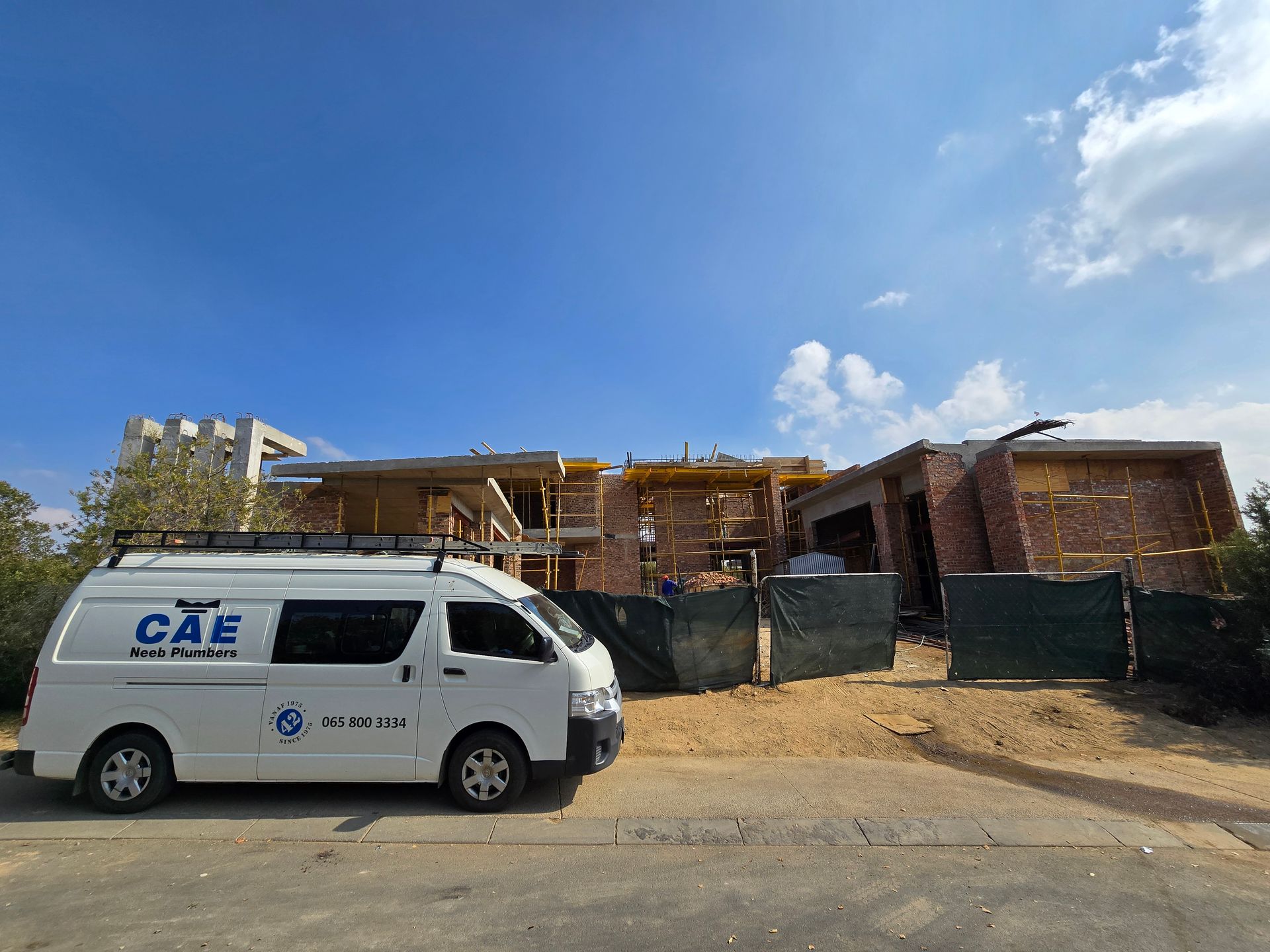 White van parked by a fenced construction site with a partly built brick house under a blue sky.