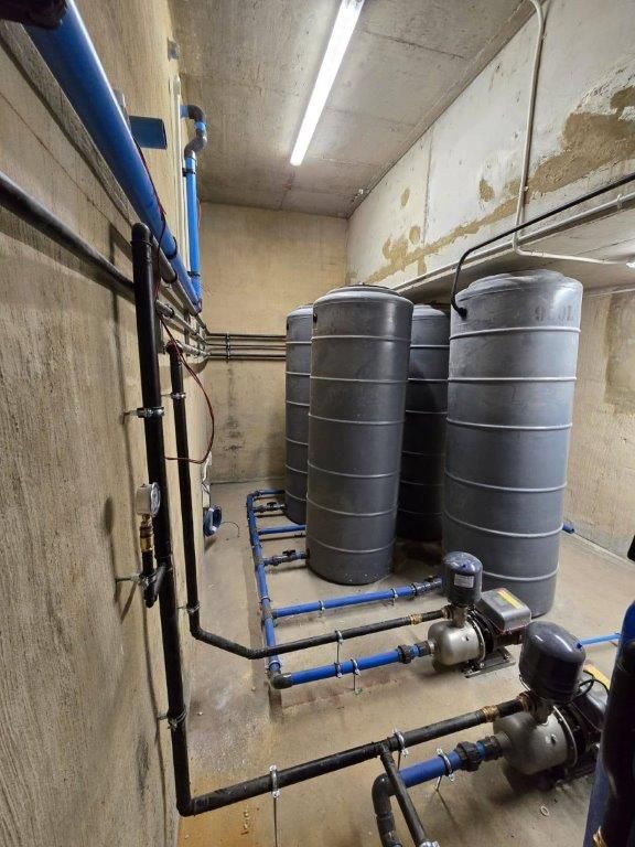 Industrial basement with gray water tanks, blue pipes, and pumps along concrete walls.