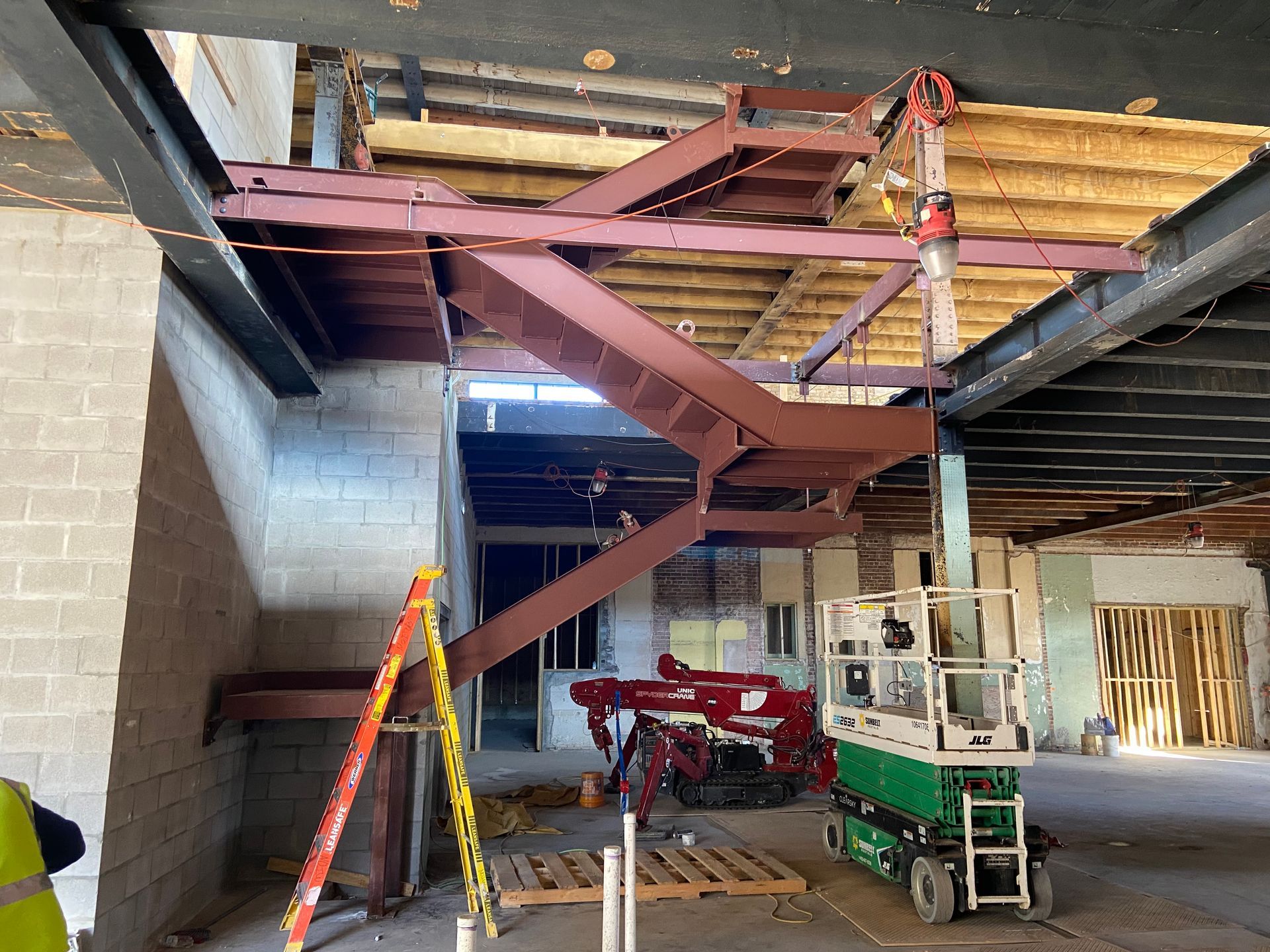 Construction site showing a metal staircase being installed, suspended by a crane inside an unfinished building.
