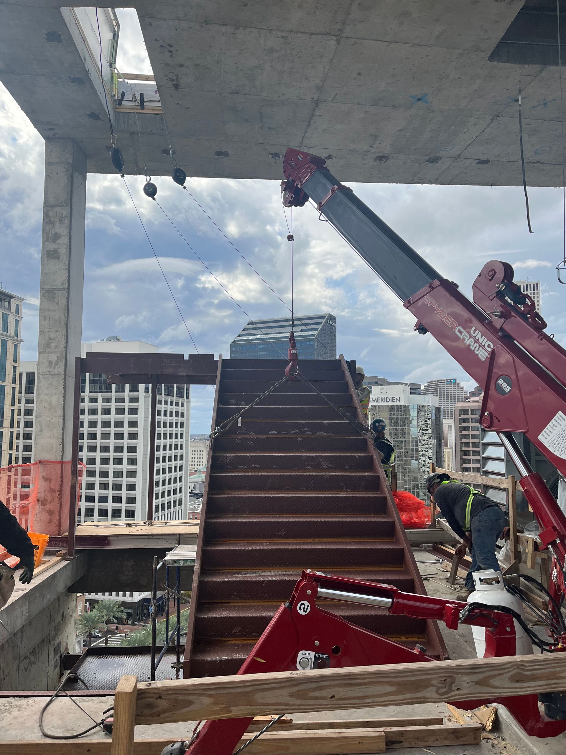 A red construction crane lifts a set of metal stairs into place on an open-floor building under construction.