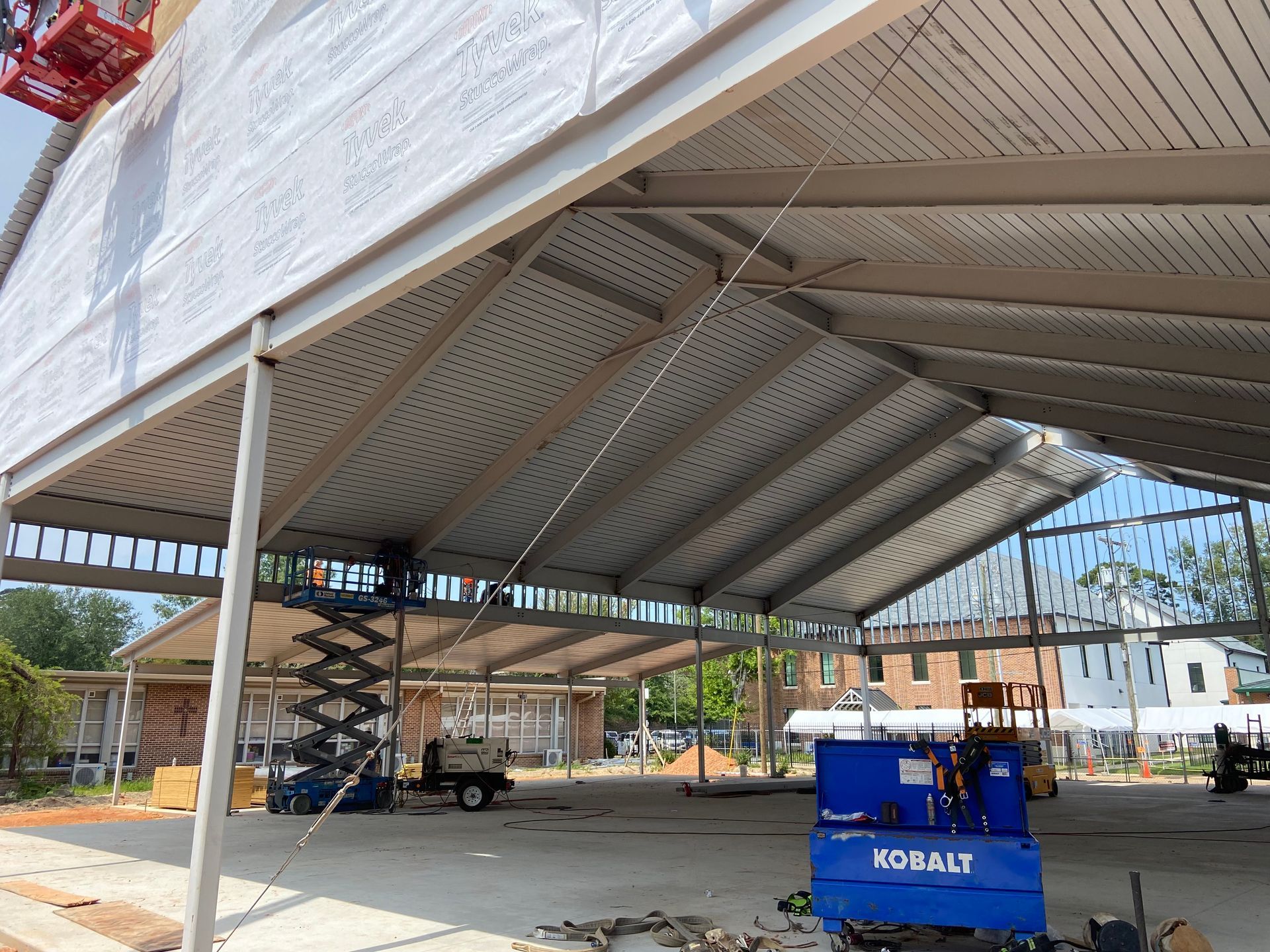 A steel-framed open-air building structure under construction, featuring a metal roof, heavy beams, and heavy equipment.