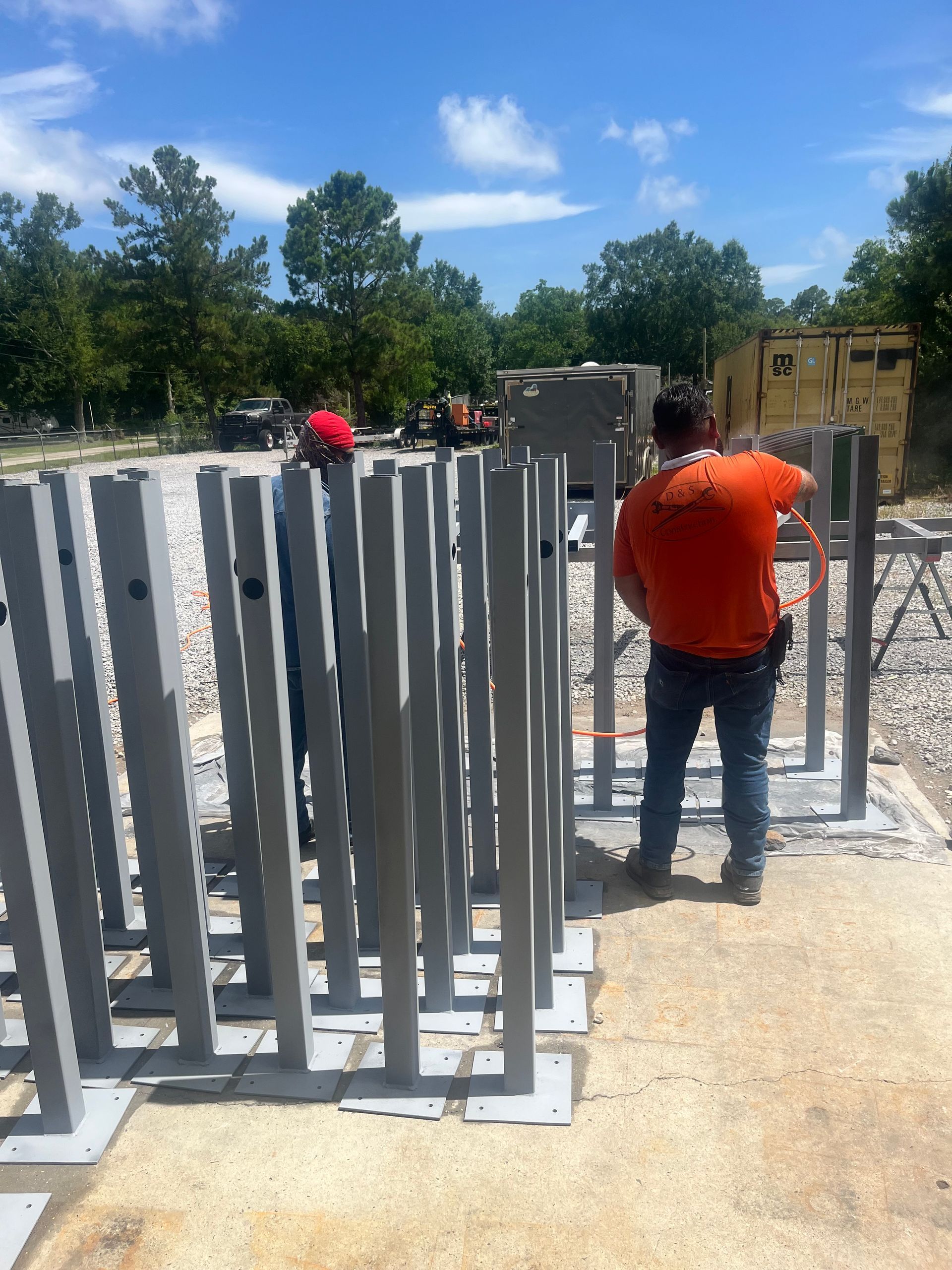 Two workers stand near rows of grey, vertical metal posts installed on a gravel construction site under a blue sky.