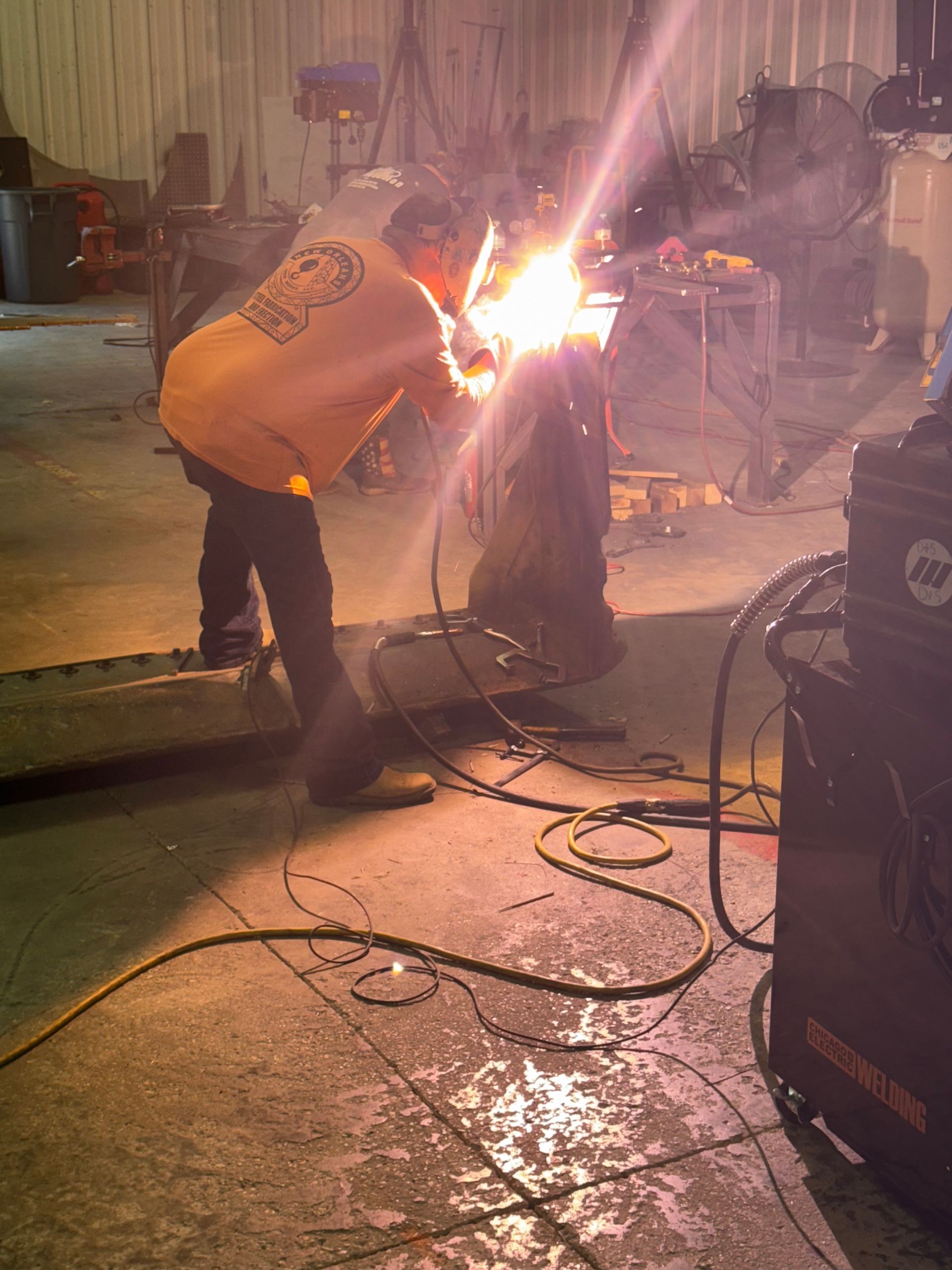 A person in an orange sweatshirt uses a welding torch, creating bright light and sparks in a workshop.