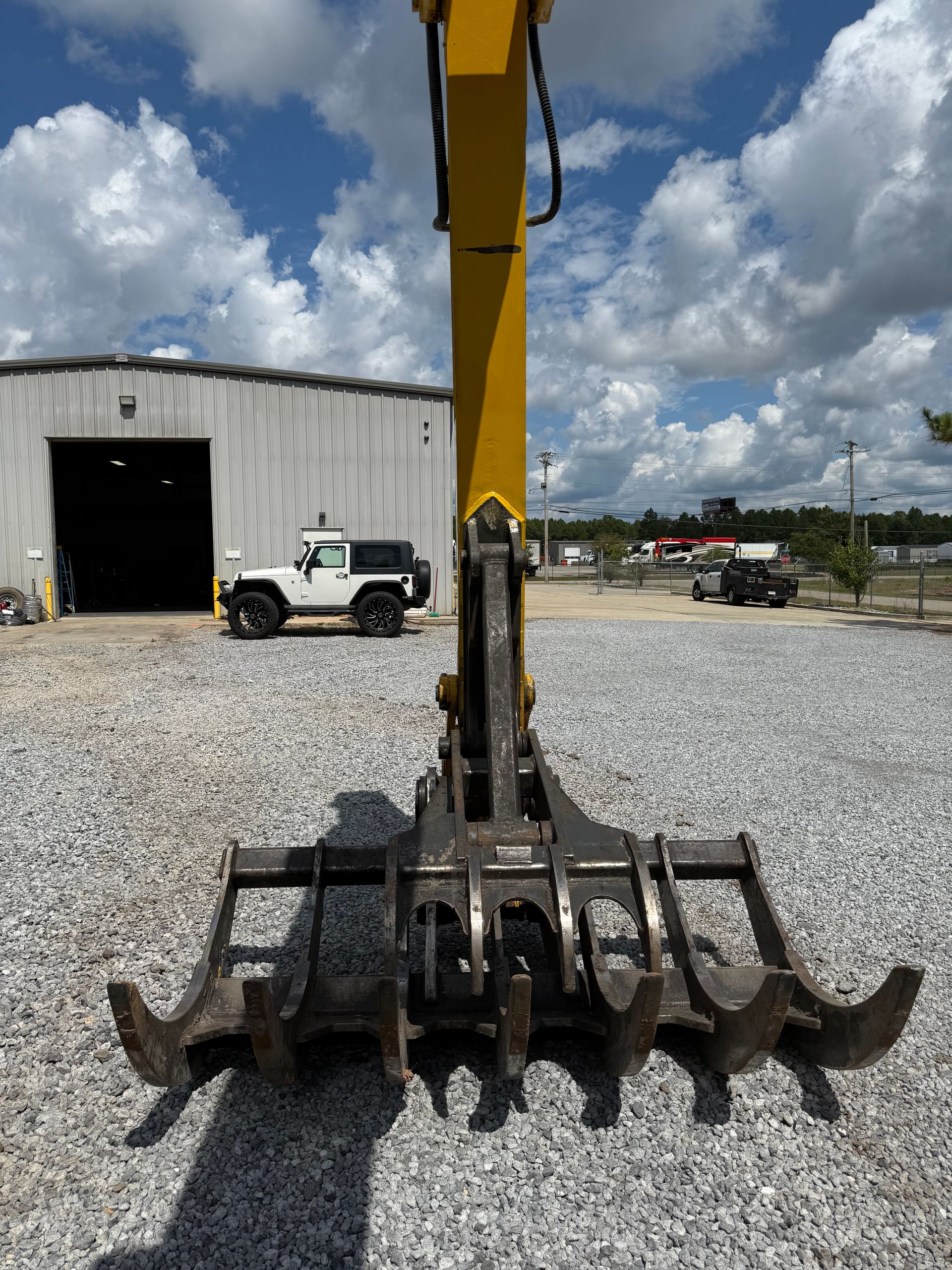 A yellow excavator boom with a large metal rake attachment parked in a gravel lot next to a white Jeep and a warehouse.