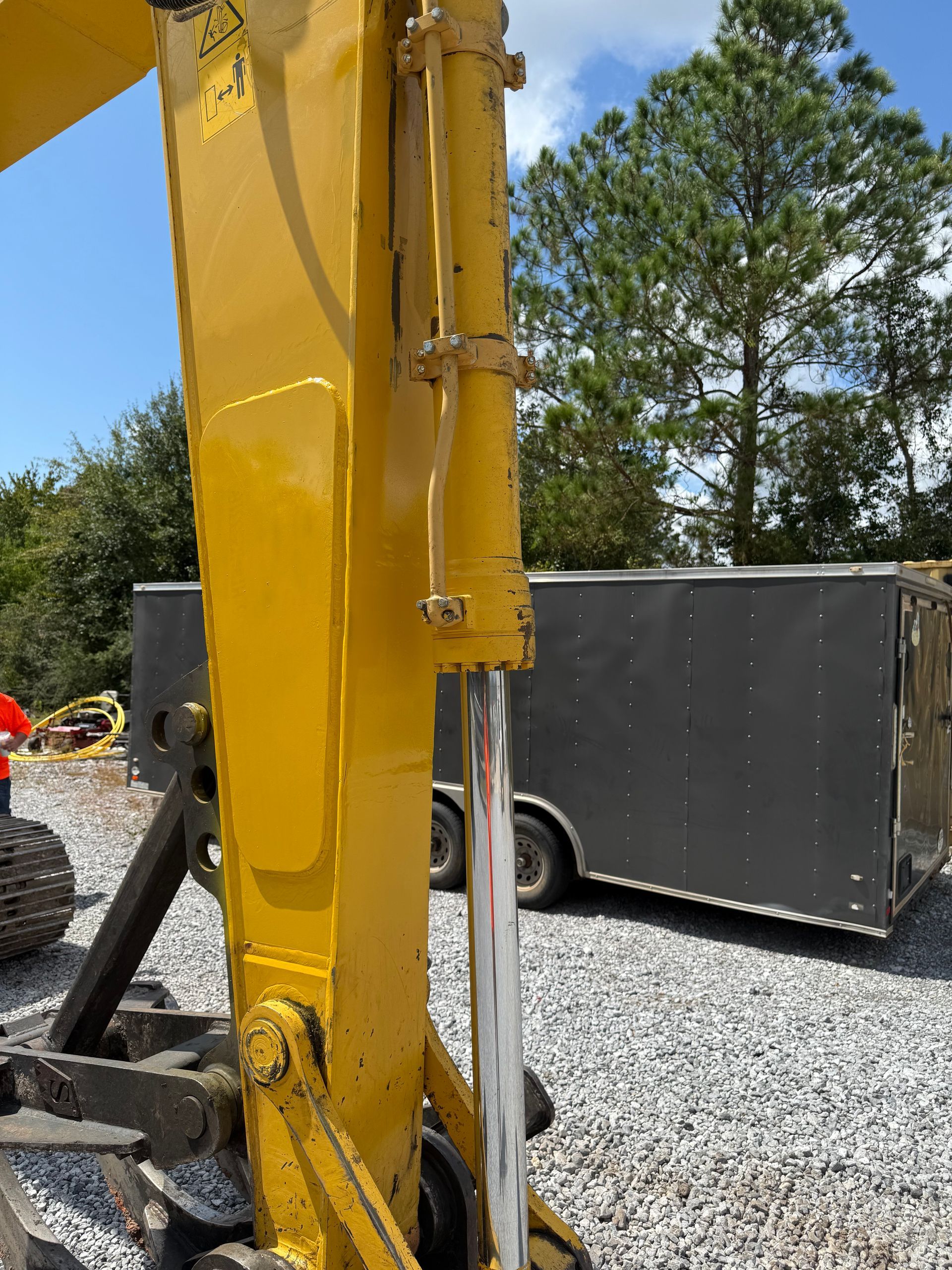 Close-up of a yellow excavator arm and hydraulic cylinder parked outdoors near a trailer on a gravel lot.