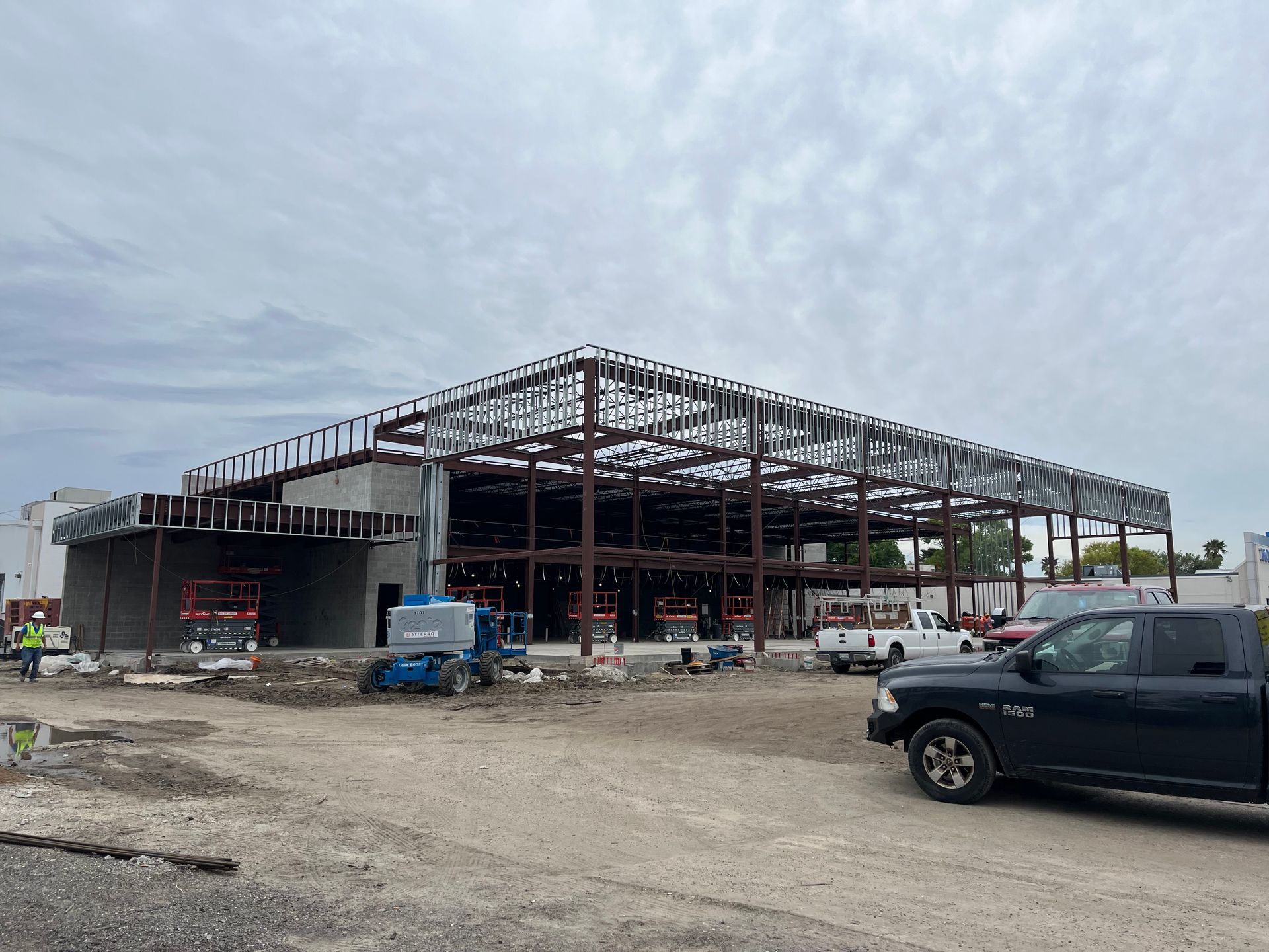 A construction site featuring an exposed steel frame building under a cloudy sky, with work vehicles in the foreground.