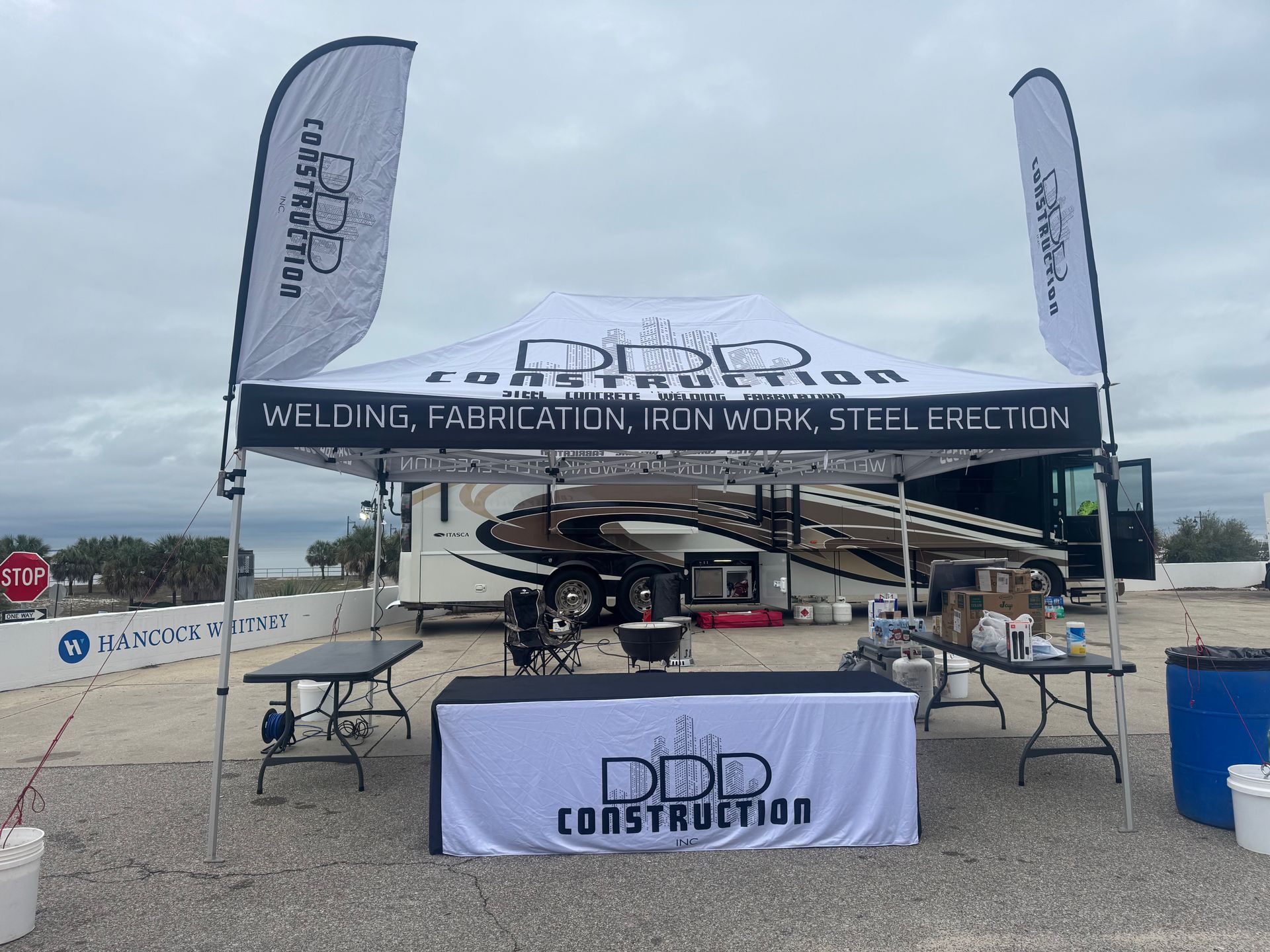 DDD Construction promotional tent with branded table and feather banners set up on a gravel lot under a cloudy sky.