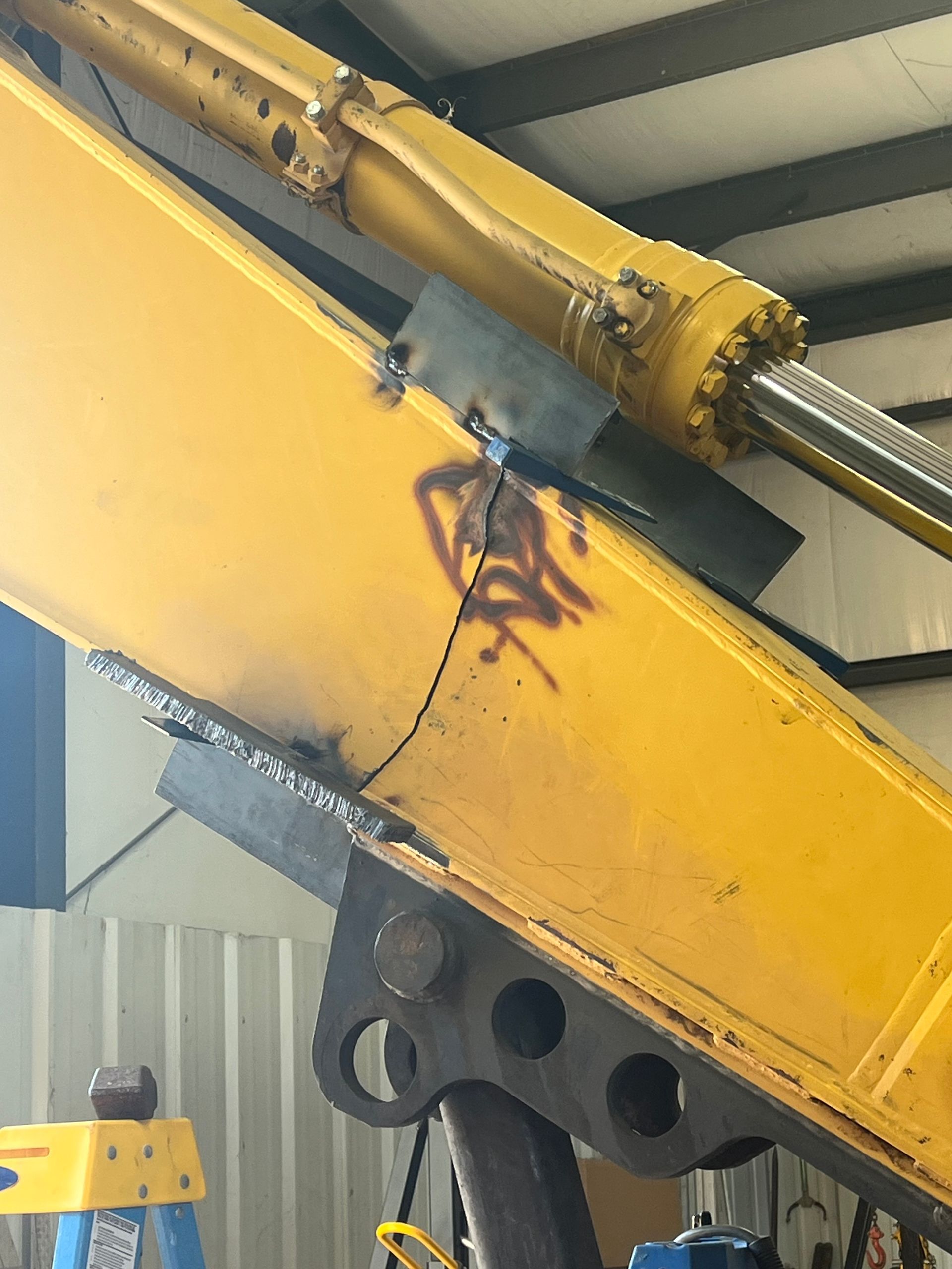 A close-up of yellow heavy equipment machinery featuring a large metal structural crack with a temporary repair plate.