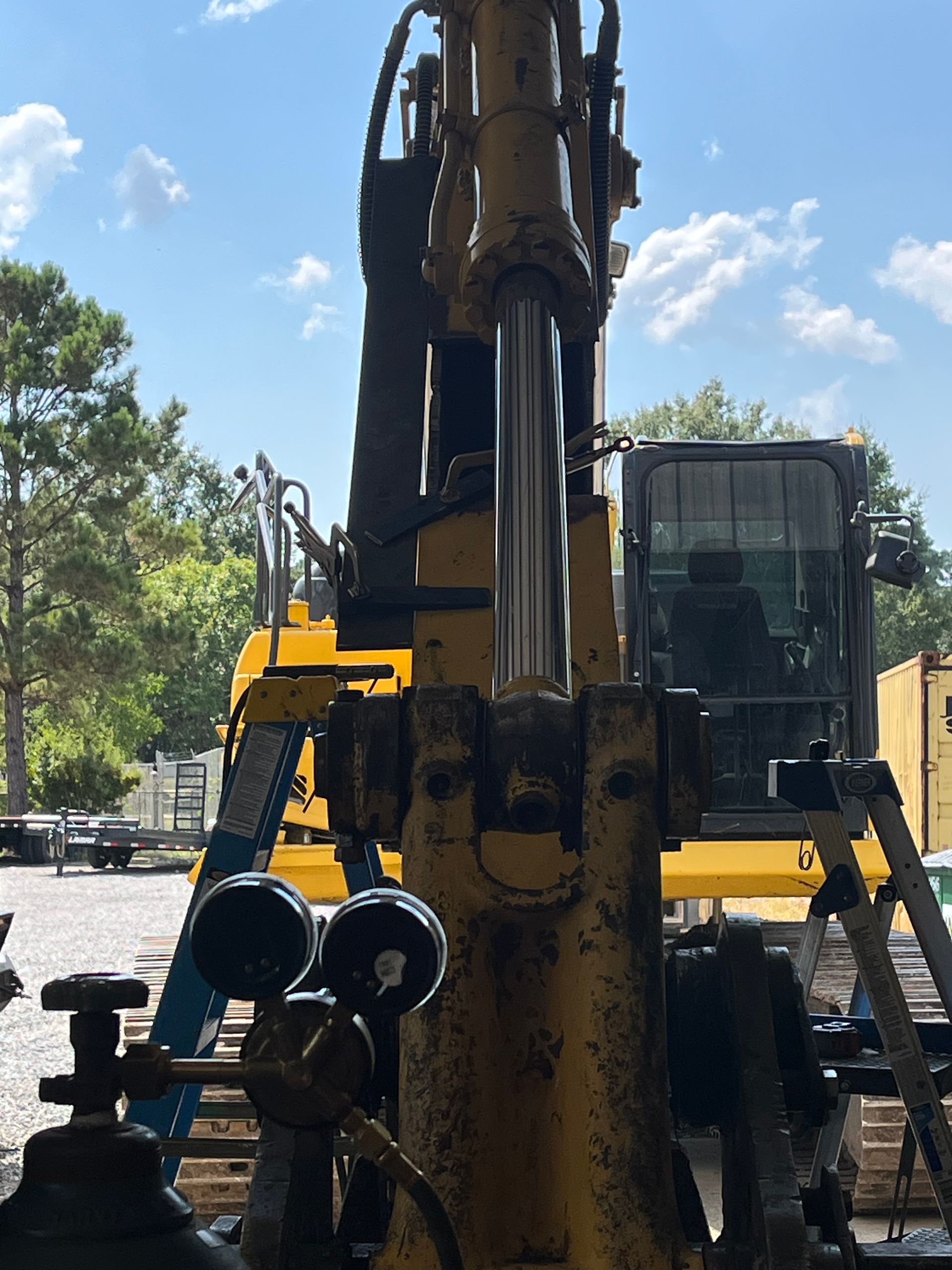 A close-up view of a yellow excavator's hydraulic arm, with a welding gas regulator in the foreground and a cabin behind.