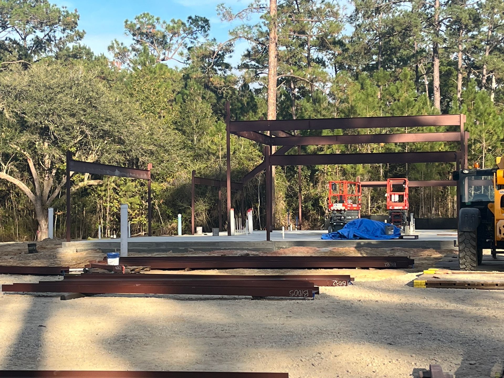 Steel beams for a building frame stand on a concrete slab at a construction site surrounded by trees.