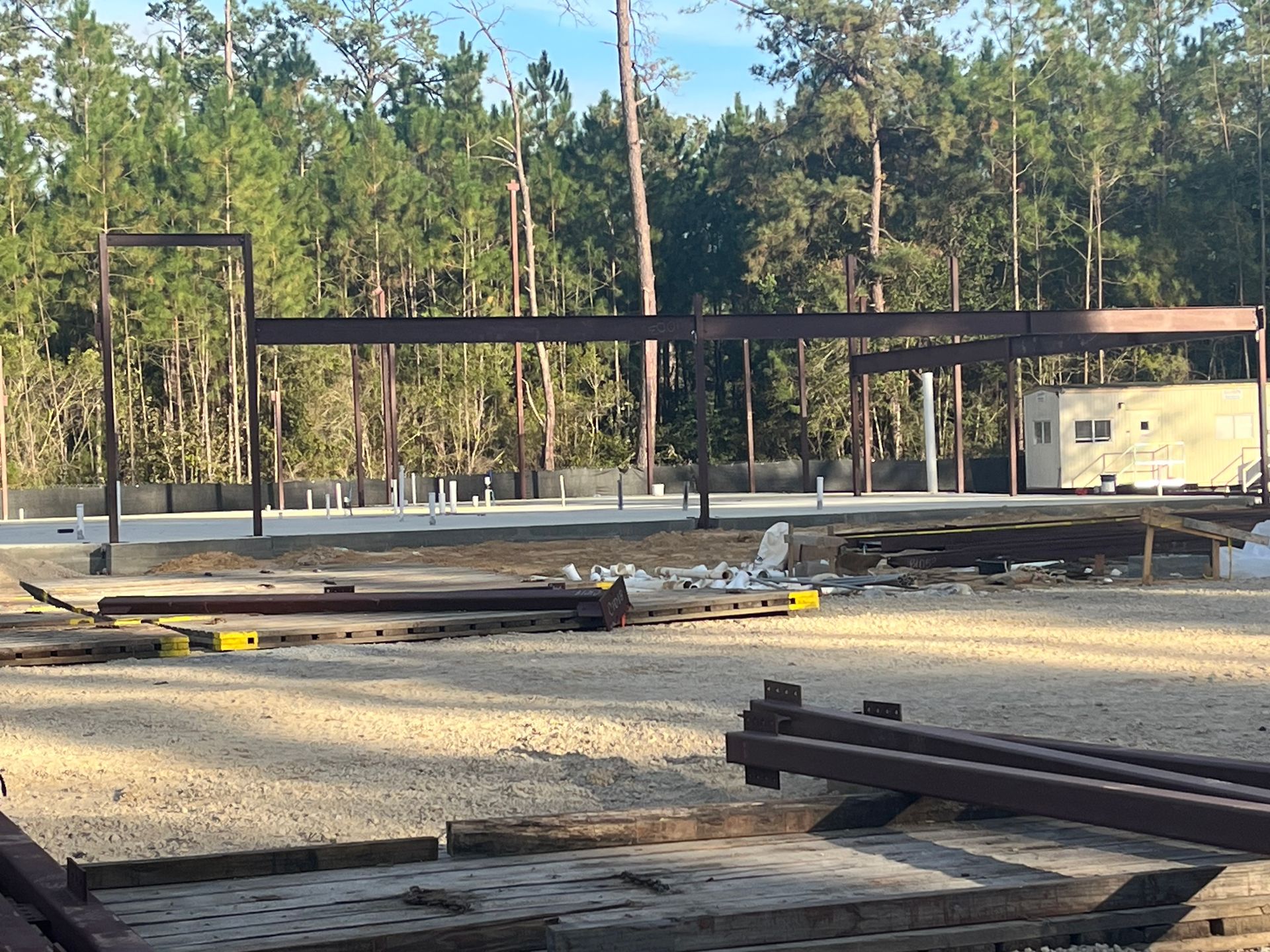 Construction site featuring steel I-beam framing over a concrete foundation in a wooded area.
