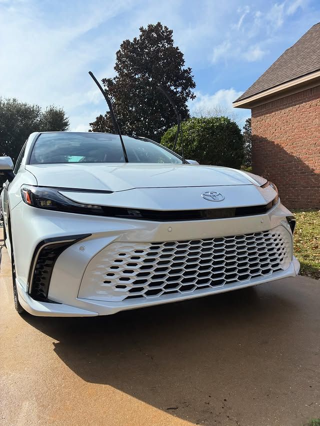White Toyota sedan parked on a sunny driveway, wipers up, in front of a house.