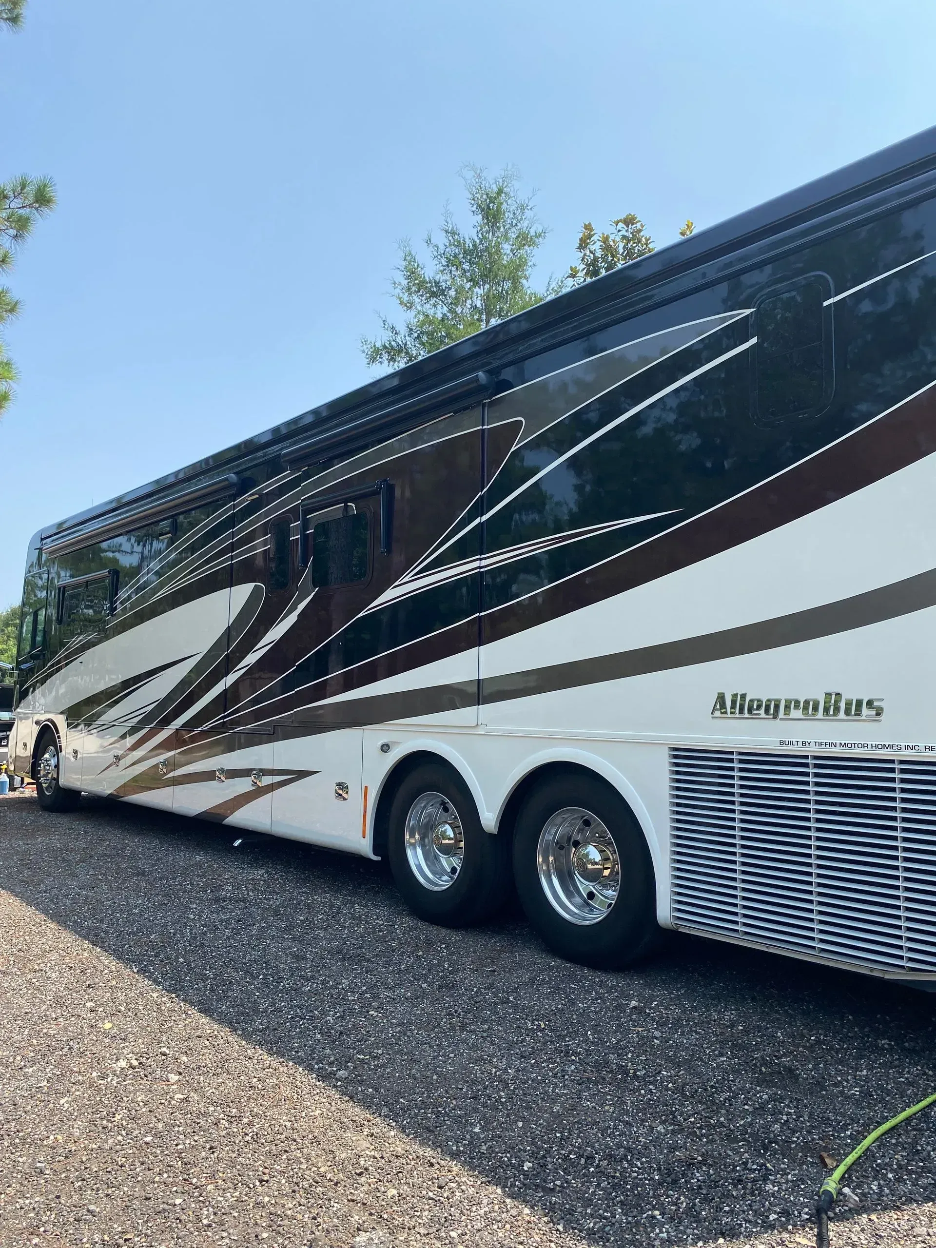 A modern, multi-axle luxury motorhome parked on a gravel surface under a clear blue sky.