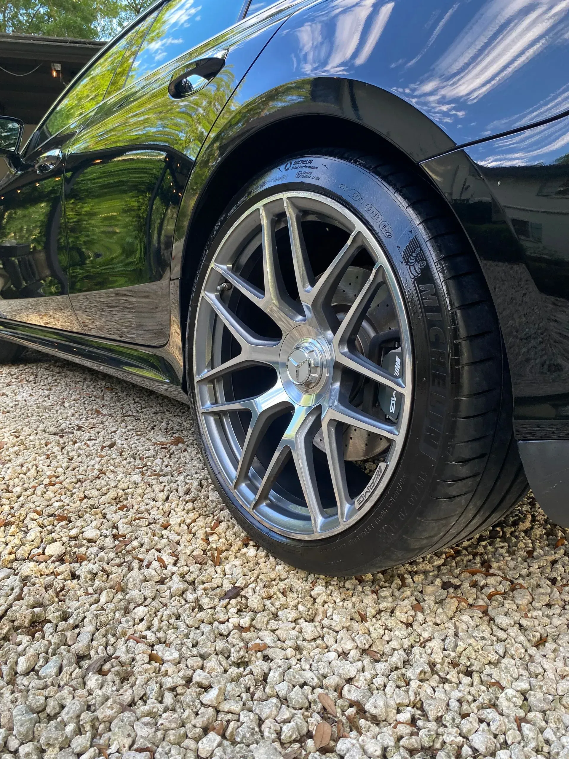 Close-up of a glossy black car's rear tire and multi-spoke metallic wheel parked on a gravel driveway.