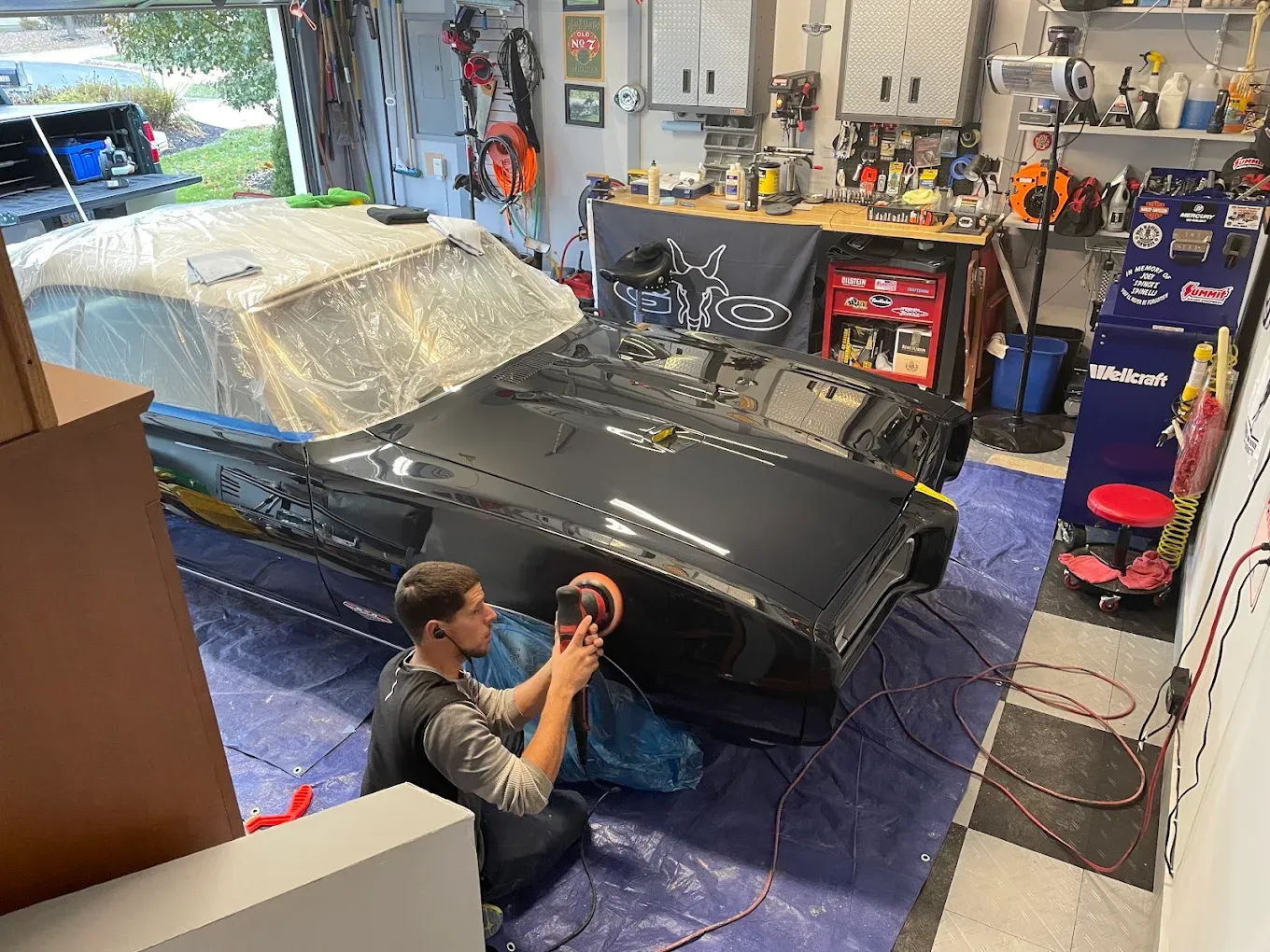 Man polishing black car panel in a garage, a covered vehicle in the background.