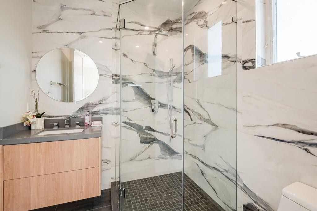 Bathroom with marble walls and glass shower door, wooden vanity, and round mirror.