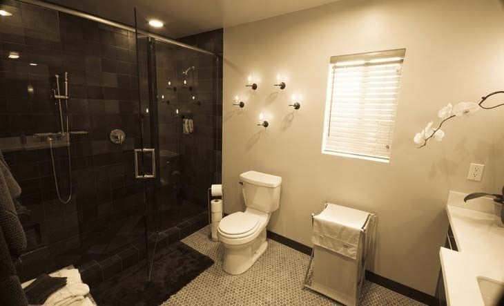 Modern bathroom with glass shower, toilet, window with blinds, and decorative sconces.