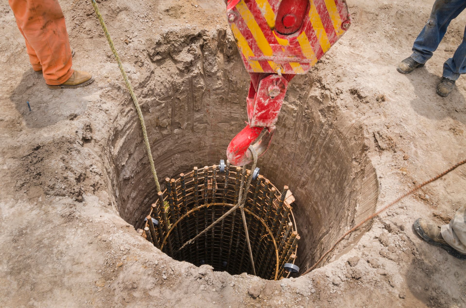 Workers lowering rebar cage into a deep, circular hole with a crane.