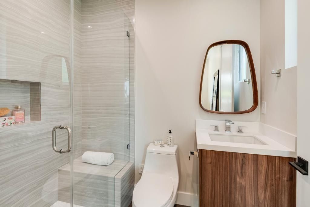 Modern bathroom with a glass shower, toilet, and wooden vanity with a unique mirror.