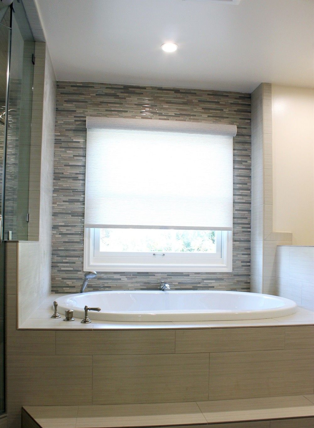 Bathroom with oval bathtub beneath a window with white shade; textured stone tile accent wall.