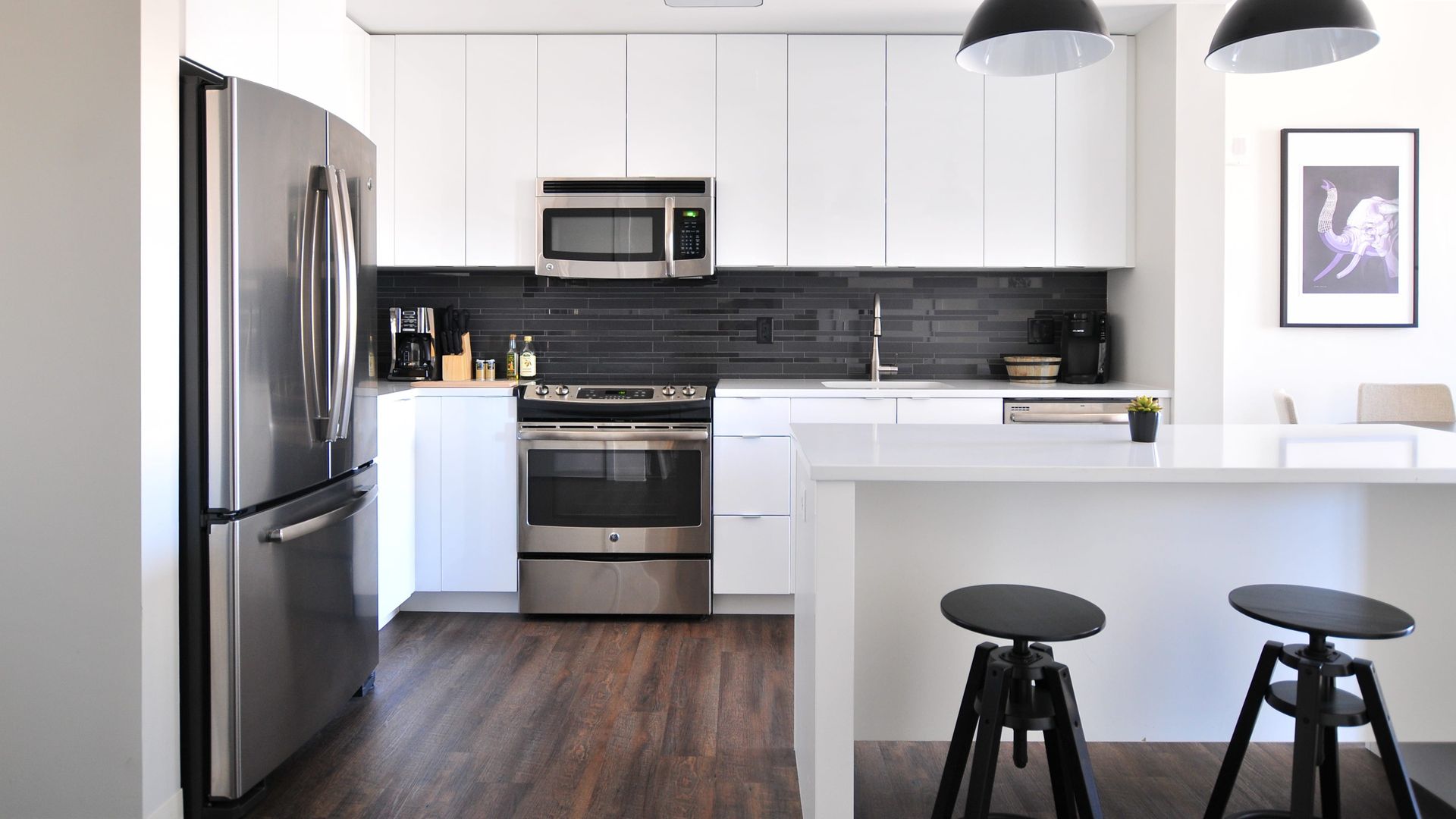 Modern white kitchen with stainless steel appliances, dark backsplash, and stools at a breakfast bar.