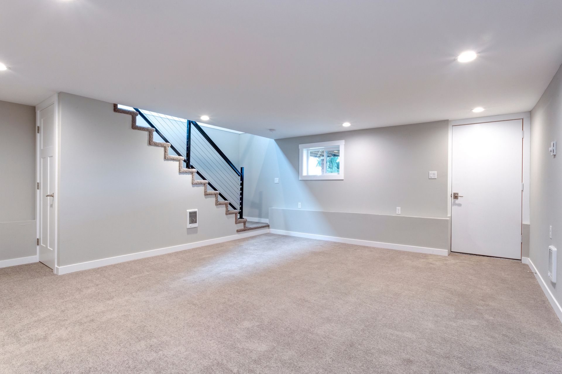 Empty finished basement with gray walls, carpet, stairs, and a white door.