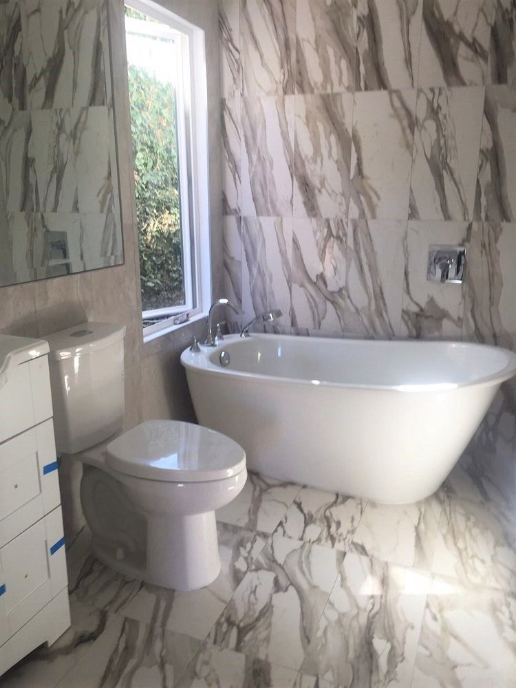 Bathroom with marble-patterned walls and floor. Features a white freestanding tub, toilet, and window with greenery.