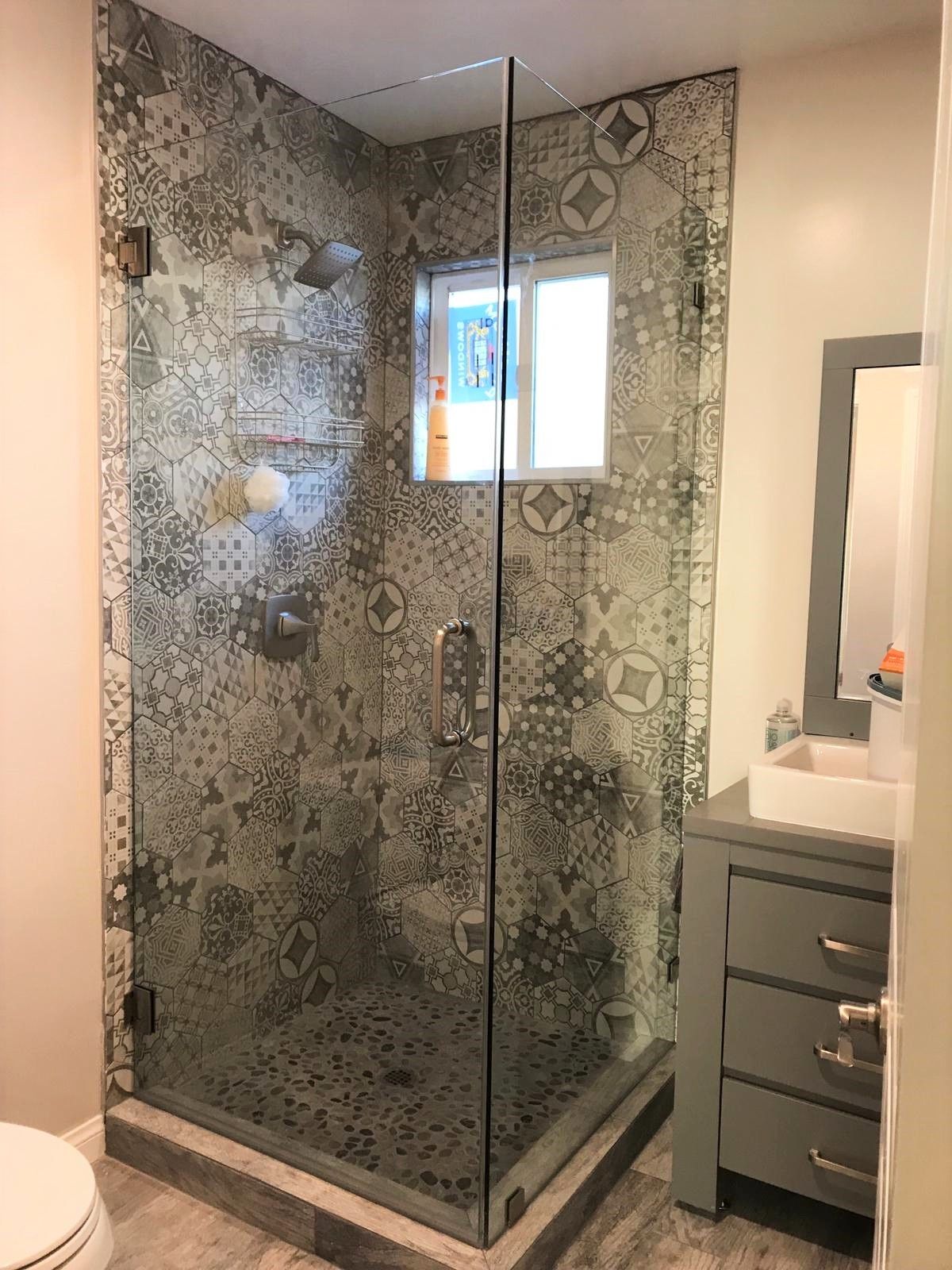 Shower with patterned gray tiles and glass door next to a gray vanity in a modern bathroom.