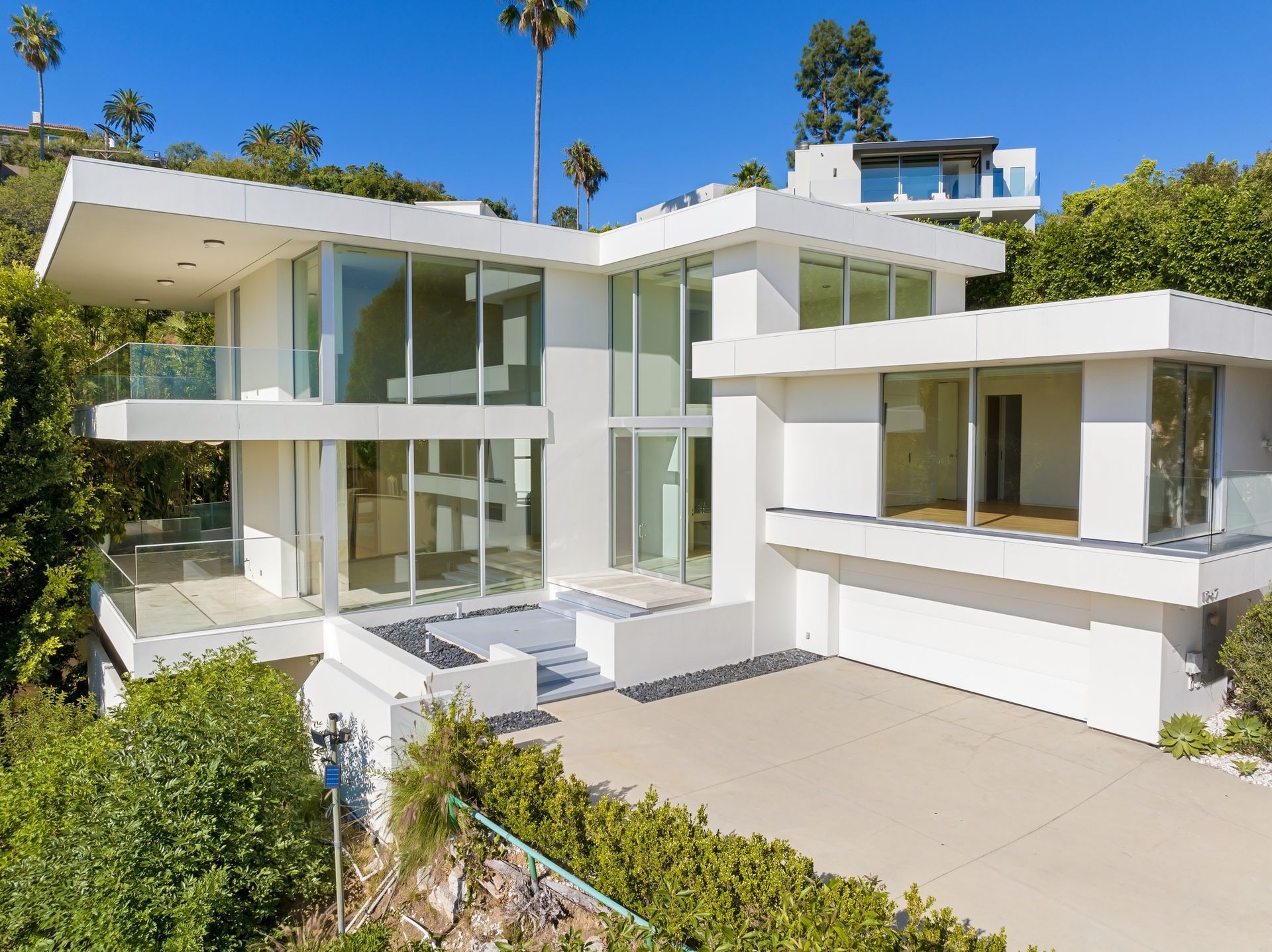 Modern, white two-story house with large glass windows and balconies, set in a hillside with palm trees.