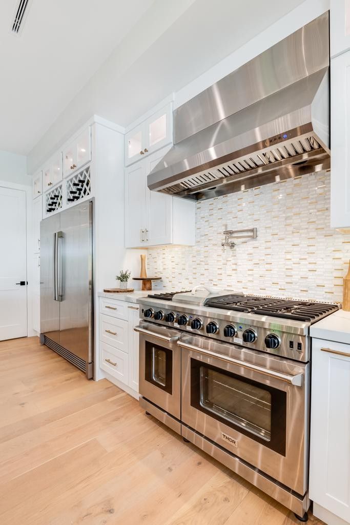 Modern white kitchen with stainless steel appliances, stove, and range hood. Light wood flooring.