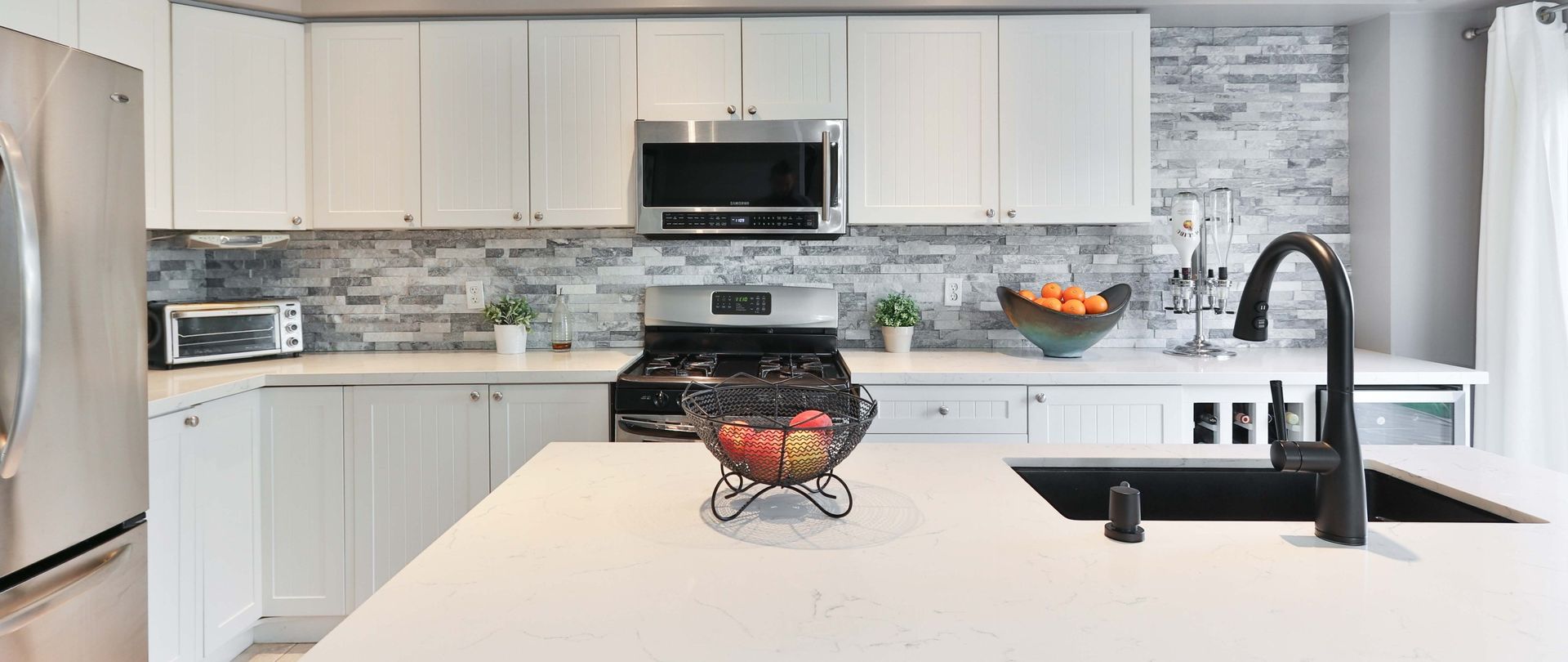 Modern white kitchen with stainless steel appliances, brick backsplash, and a black faucet over a sink.