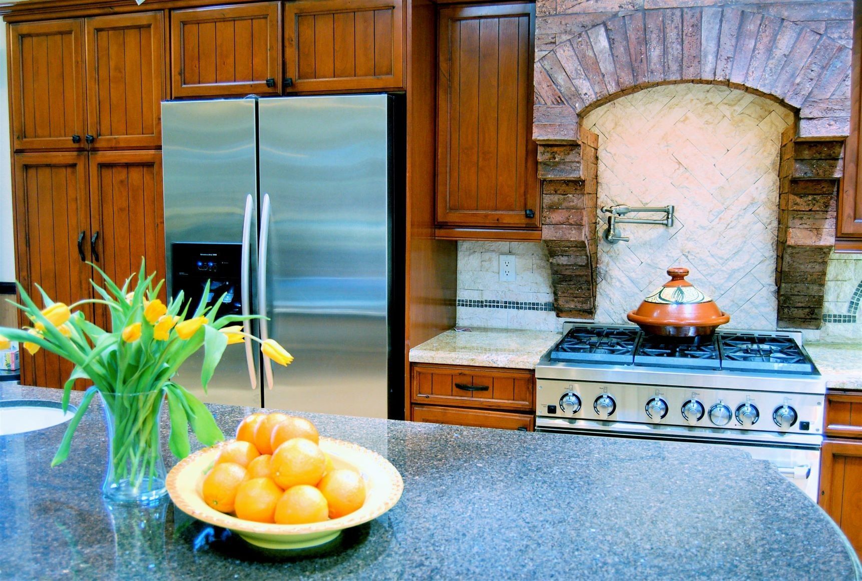 Kitchen with wooden cabinets, stainless steel fridge, brick arch, granite countertop, and flowers.