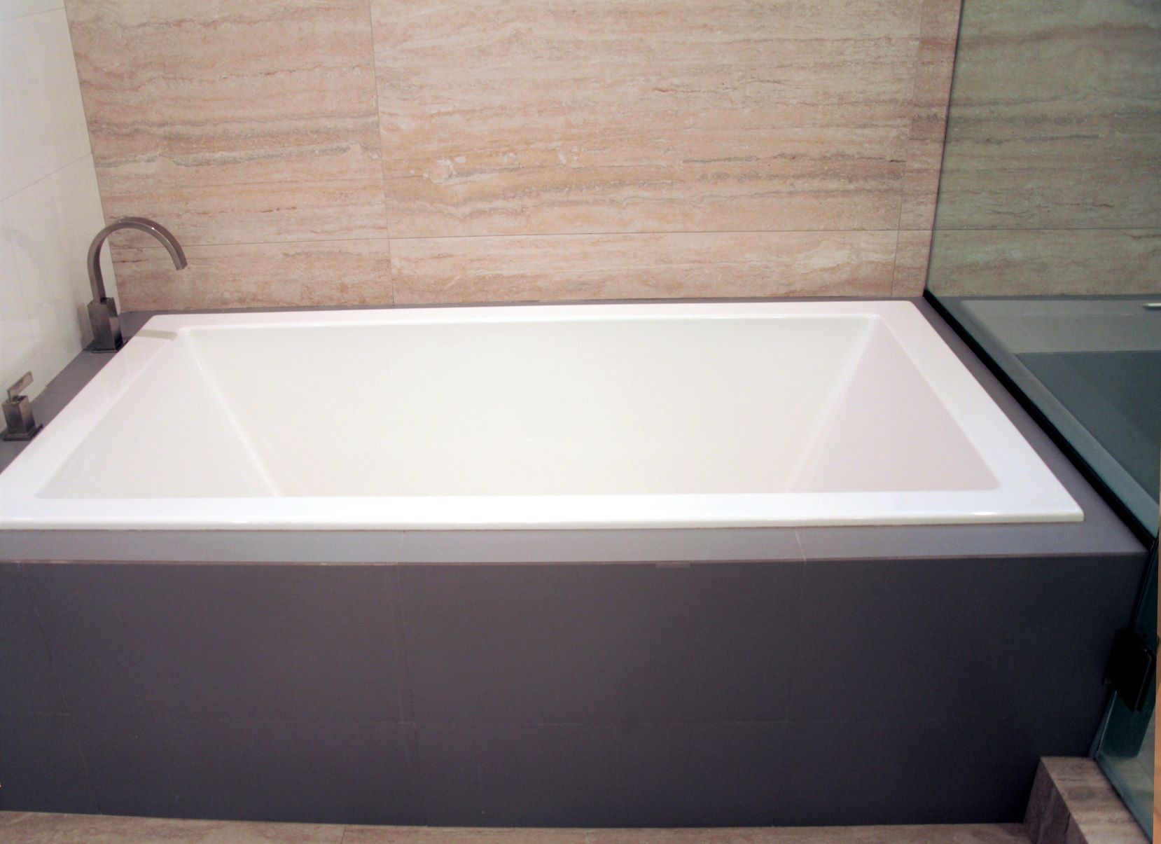 White rectangular bathtub in a grey enclosure, set against a light-colored tiled wall.