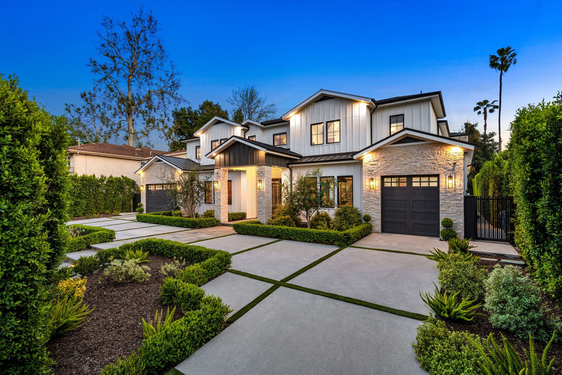 Modern two-story house with a stone and white exterior, lush landscaping, and a driveway.