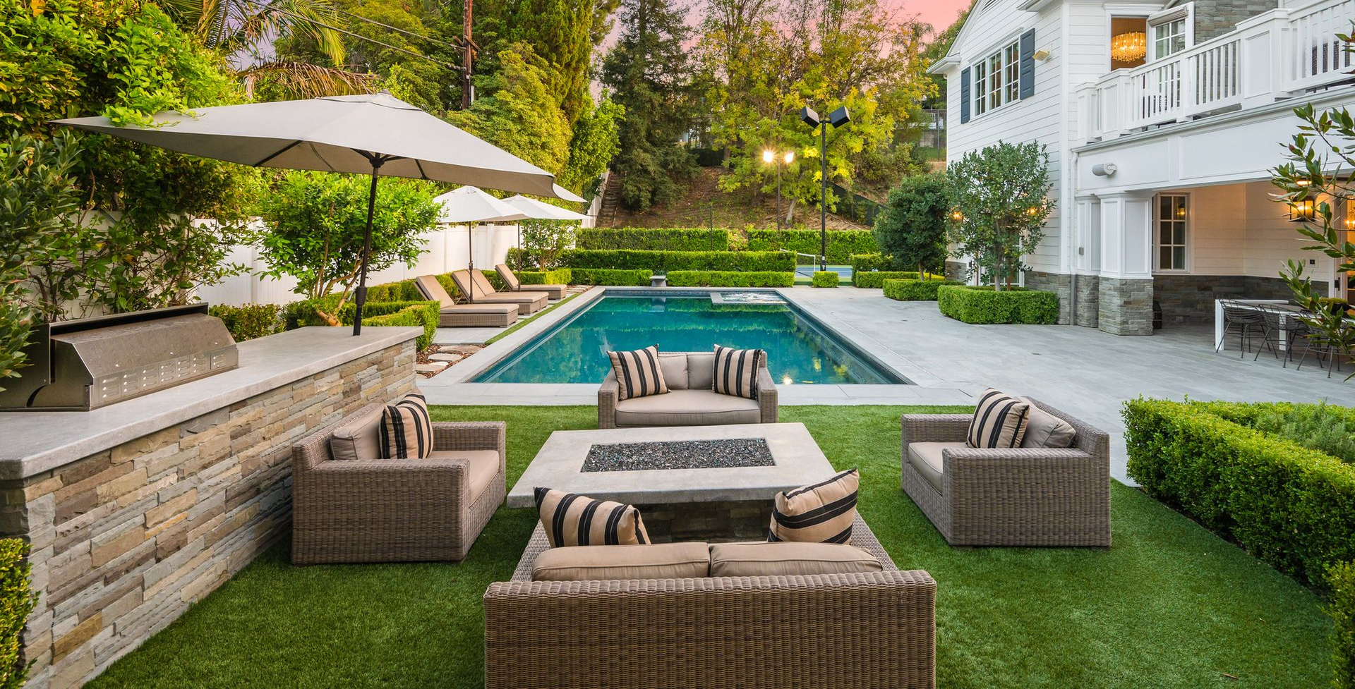 A backyard with a swimming pool, stone outdoor kitchen, and wicker seating area arranged around a fire pit.
