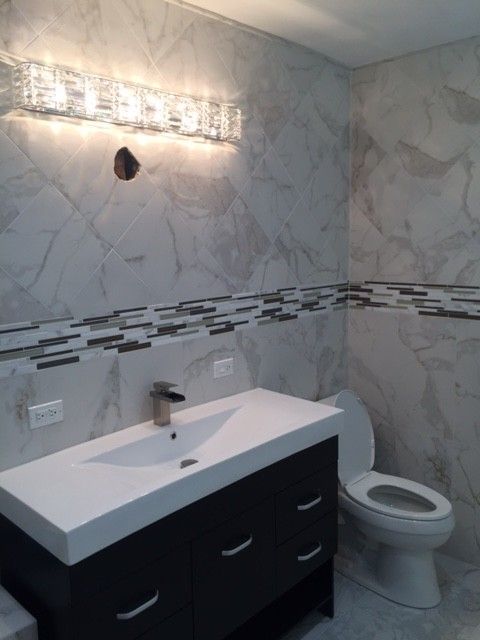 Modern bathroom with a white vanity, toilet, and marble-like tile walls. A decorative light fixture is above the vanity.