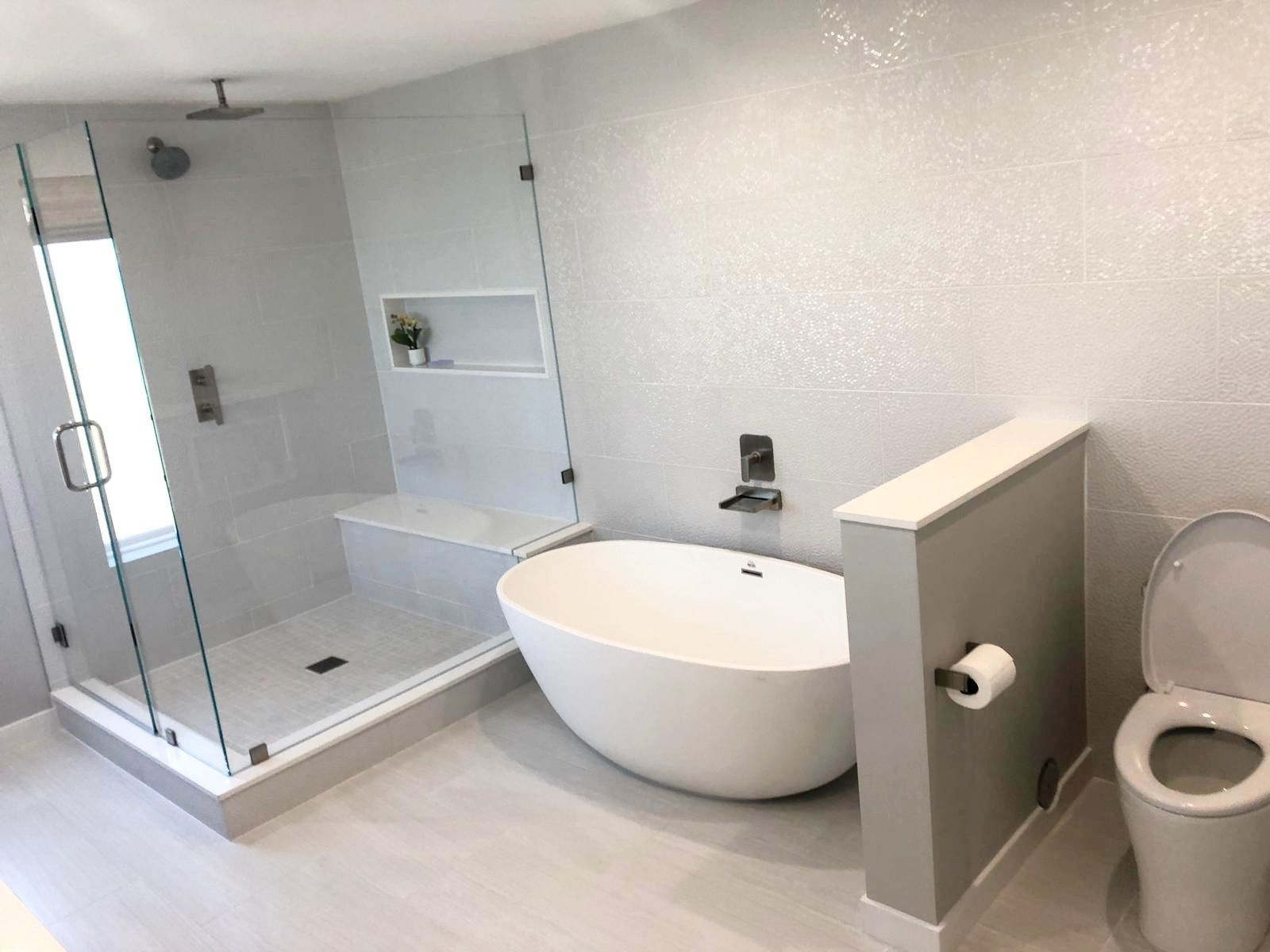 Modern bathroom with a glass shower, white tub, and toilet. Light gray walls and flooring.