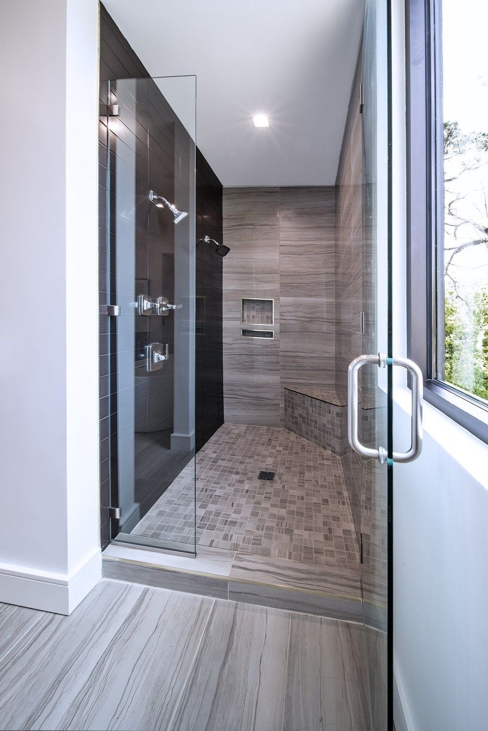 Modern shower with stone tile, glass door, bench, and window.
