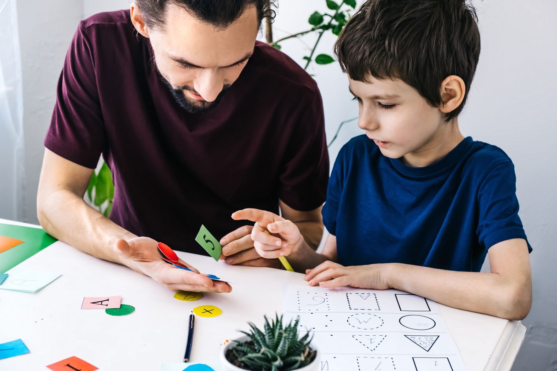 Adult helping child with craft project at a table, drawing with colorful shapes and paper.